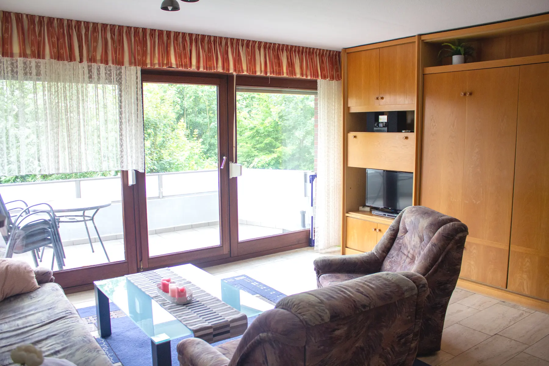 Wohnzimmer mit Balkonfenstern, Sesseln und Schrankbett